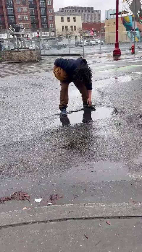 这是发生在美国街头的离谱一幕，一名露宿街头的人直接蹲在路面的积水坑旁，用积水洗脸、搓洗头发
